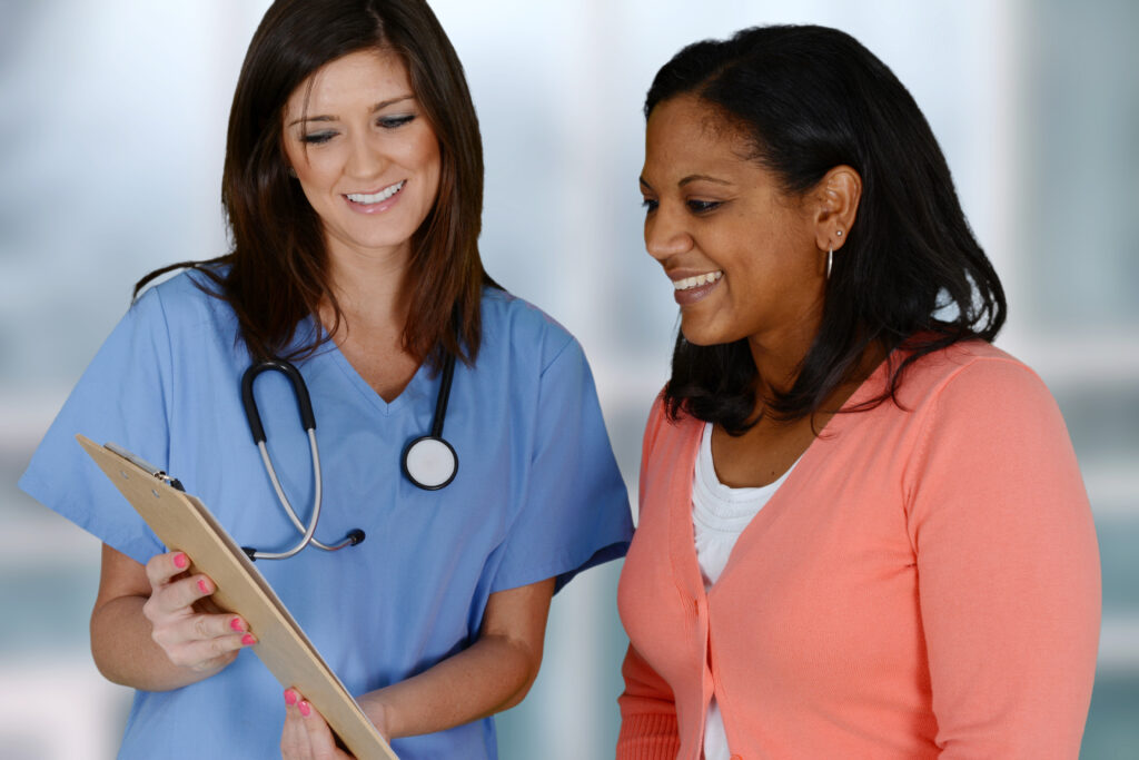 A woman in an orange shirt smiling while looking at a medical chart, held by a brunette wearing blue medical scrubs.